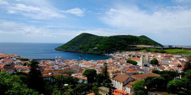 Coastal town with red-roofed buildings, lush green hills, and ocean view under a blue sky.