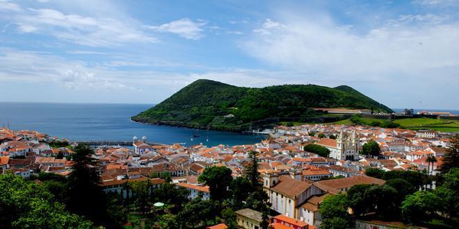 Coastal town with red-roofed buildings, lush green hills, and ocean view under a blue sky.
