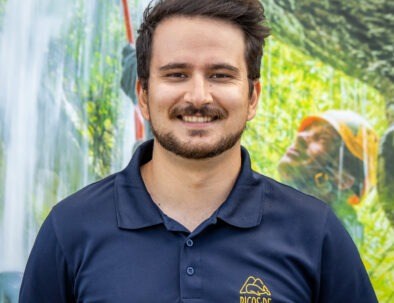 Smiling man in navy shirt stands against outdoor backdrop of trees and a person in helmet.