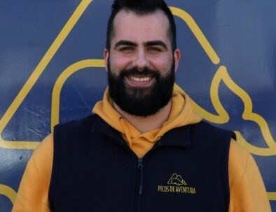 Smiling man in yellow hoodie and vest with adventure logo on blue backdrop.