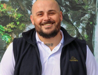 Man smiling in a vest and shirt, standing outdoors with greenery behind.