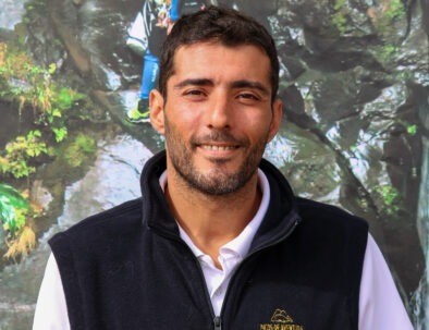 Smiling man wearing a black vest in front of a rocky, leafy background.