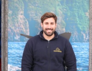 Man in black jacket standing in front of ocean and cliff backdrop.