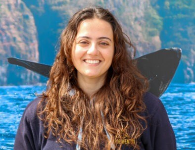 Woman with long hair smiling, whale tail and rocky cliffs in the background.