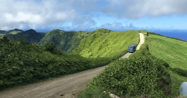 Blue van on a dirt road winding through green hills under a cloudy sky.