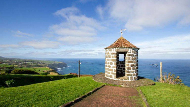 Stone gazebo on grassy hill overlooking ocean and coastline.