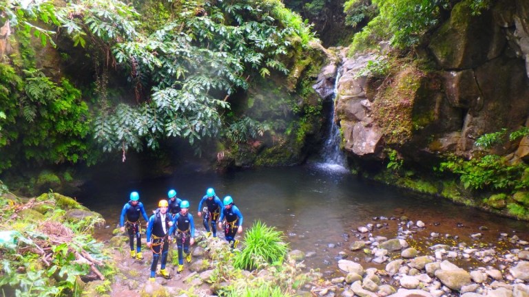 Group of people in wetsuits and helmets hiking near a forest waterfall.