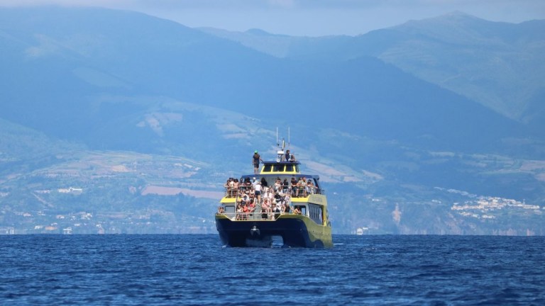 Ferry with people crossing a calm sea, mountains in the background.