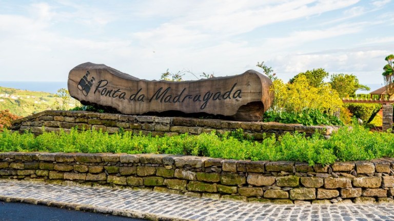 Wooden sign reading 'Ponta da Madrugada' on a stone base with greenery.