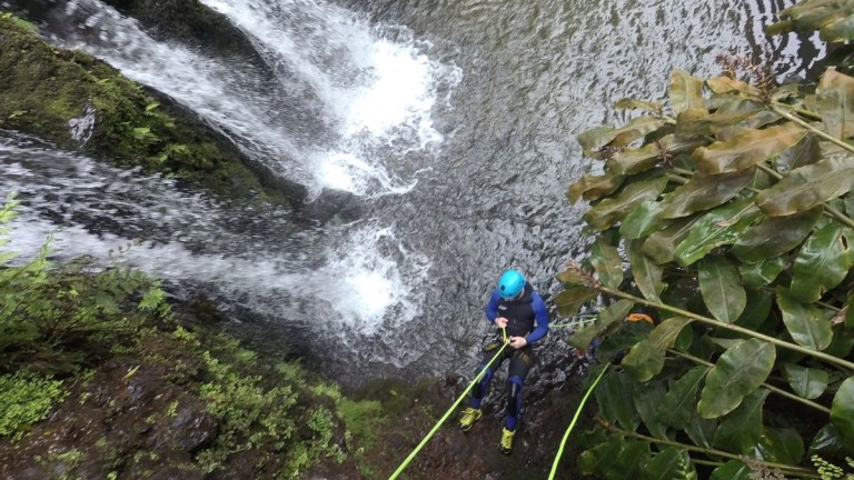 Person in blue helmet rappelling down beside a waterfall into a river.