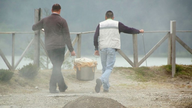Two people carrying a large pot with steam rising, near a wooden fence outdoors.