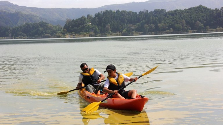 Two people kayaking on a calm lake with forested hills in the background.