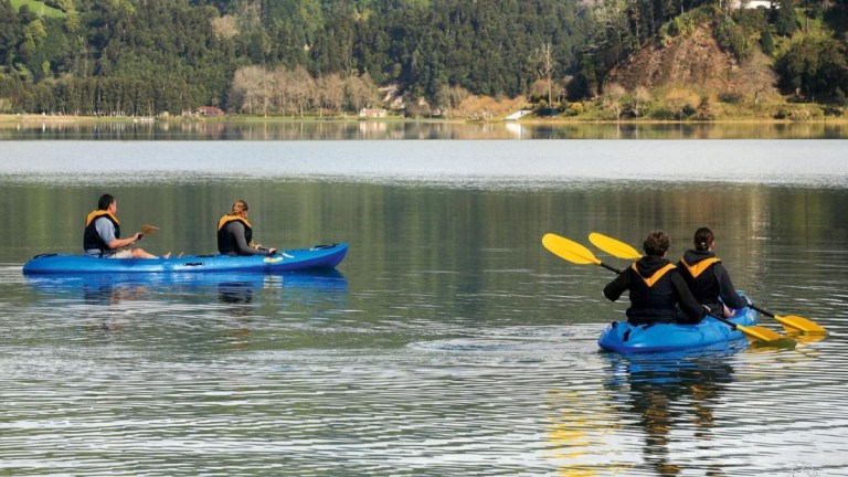 Three people kayaking on a calm lake with forested hills in the background.