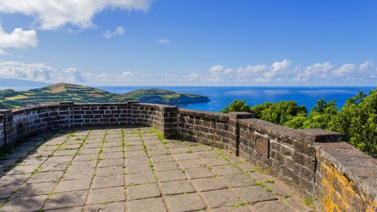 Stone terrace overlooking ocean and hilly landscape under a blue sky.