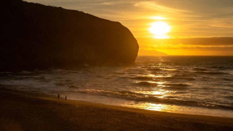 Sunset over ocean with waves, large cliff silhouette, and two figures on the beach.