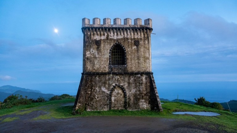 Stone tower on grassy hill at dusk with moon in cloudy sky.