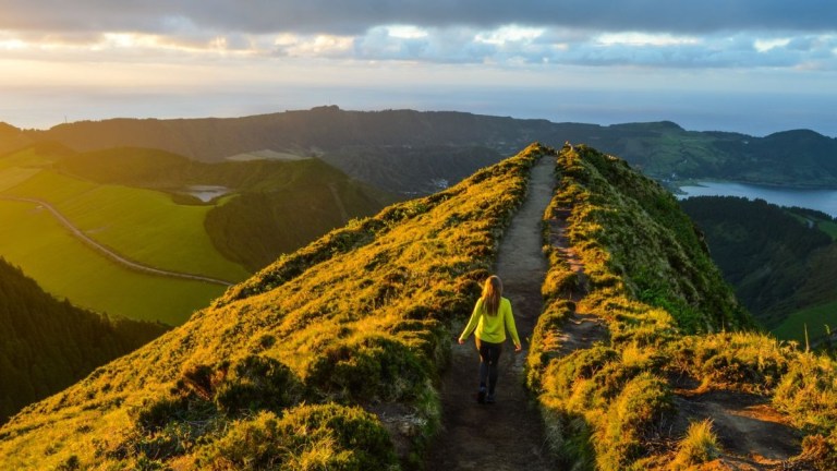 Person walking on a grassy ridge at sunset with distant hills and ocean view.