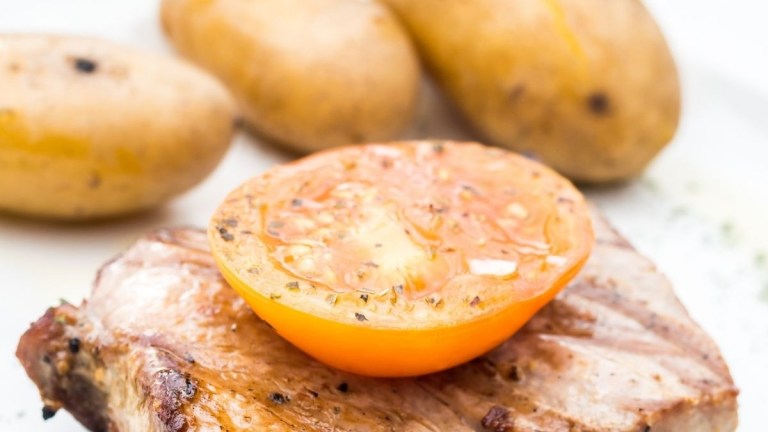 Grilled meat topped with tomato slice, and potatoes in background.