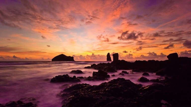 Colorful sunset over rocky coastline with silhouetted island.