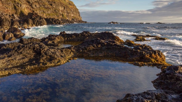 Rocky coastline with tide pools and ocean waves under a partly cloudy sky.
