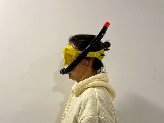 Person in profile wearing snorkel and yellow mask against a plain wall.