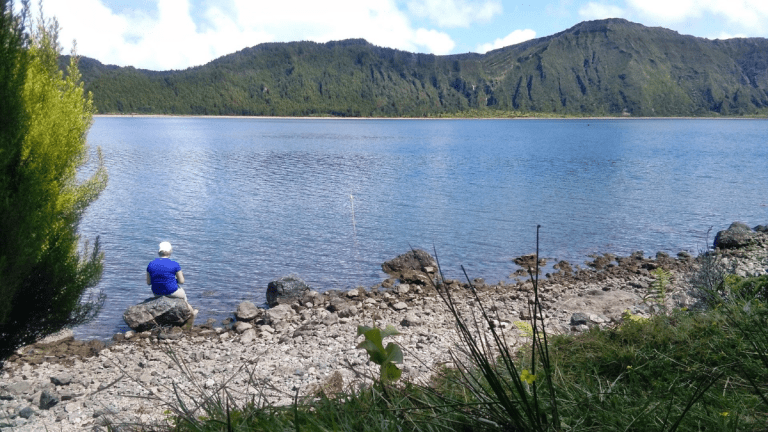 Person sits by a rocky lakeshore with mountains in the background.