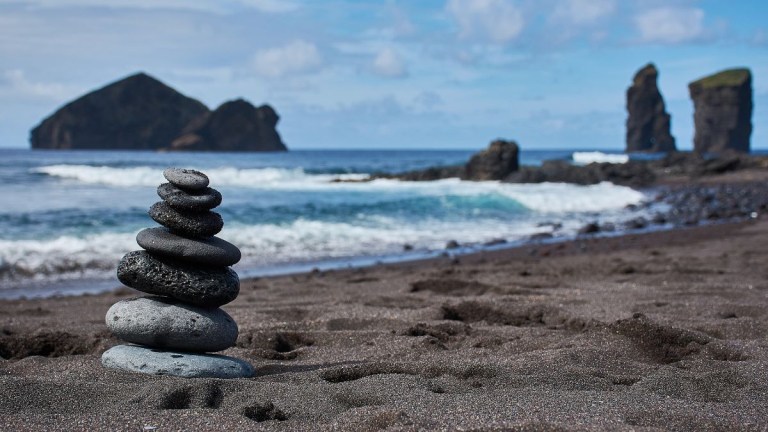 Stacked stones on a sandy beach with ocean and rock formations in the background.