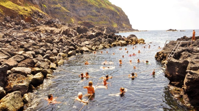 People swimming in a rocky sea inlet with hills in the background.