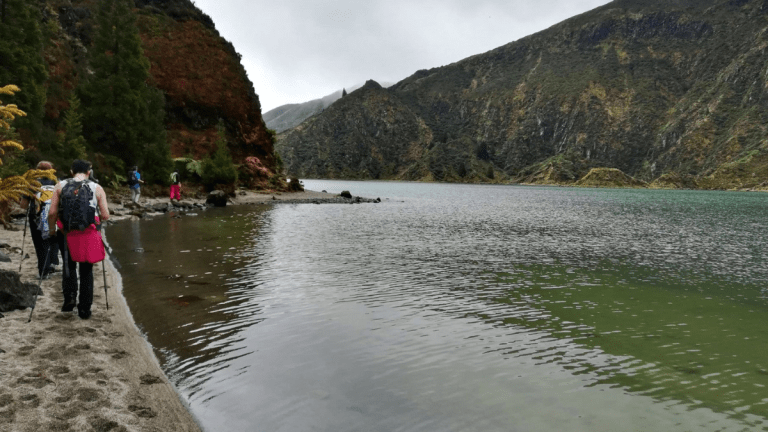 Hikers walking by a mountain lake under a cloudy sky.