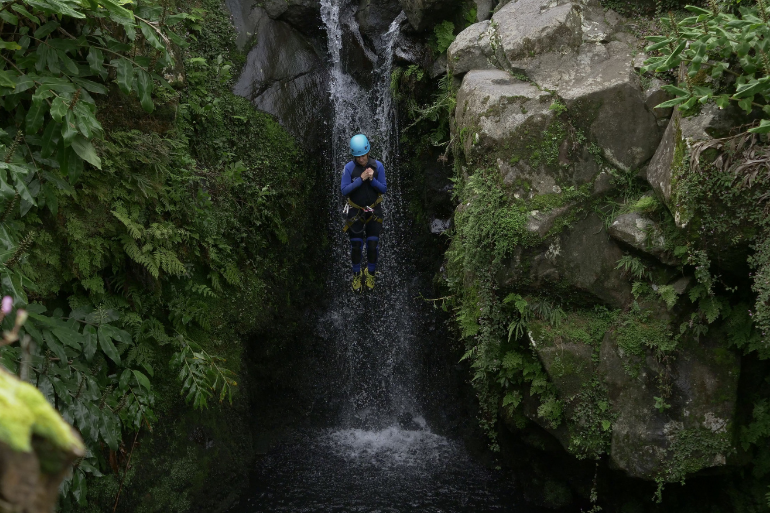 Person in helmet rappelling down a waterfall surrounded by greenery and rocks.