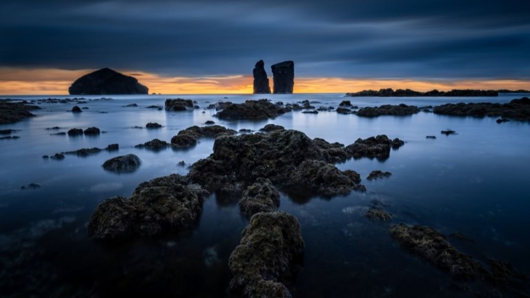 Rocky coastal scene at sunset with dark clouds and calm sea.