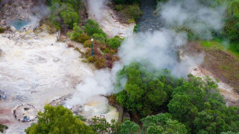 Aerial view of steaming geothermal area with trees and a river.