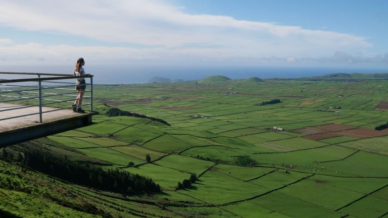 Person stands on a platform overlooking vast green fields under a blue sky.