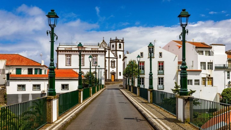 Narrow street leading to a church, flanked by white buildings and street lamps under a blue sky.