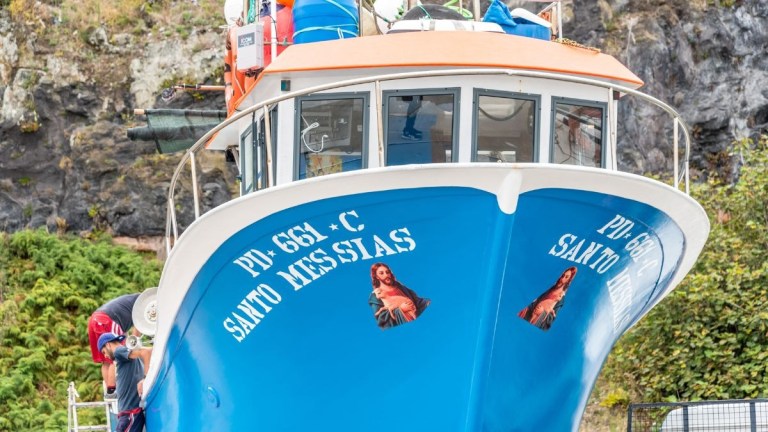 Blue and white boat with religious decals docked by a rocky shore.