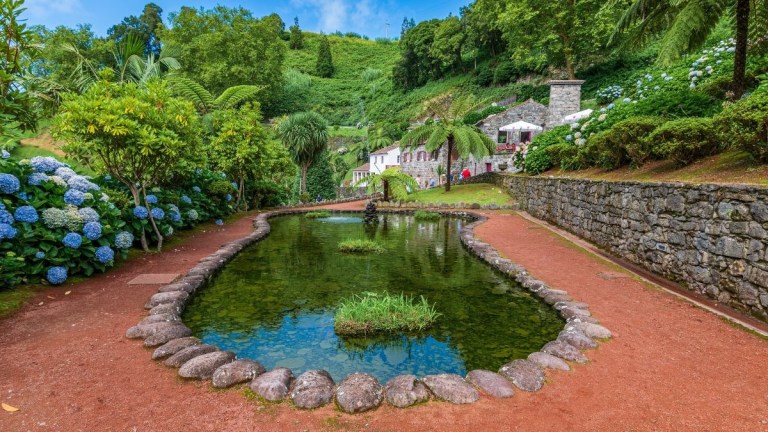 Scenic pond with stone edge, surrounded by lush greenery and flowers, with a white building in the background.