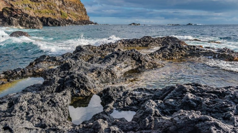 Rocky coastline with tidal pools and waves under a cloudy sky.