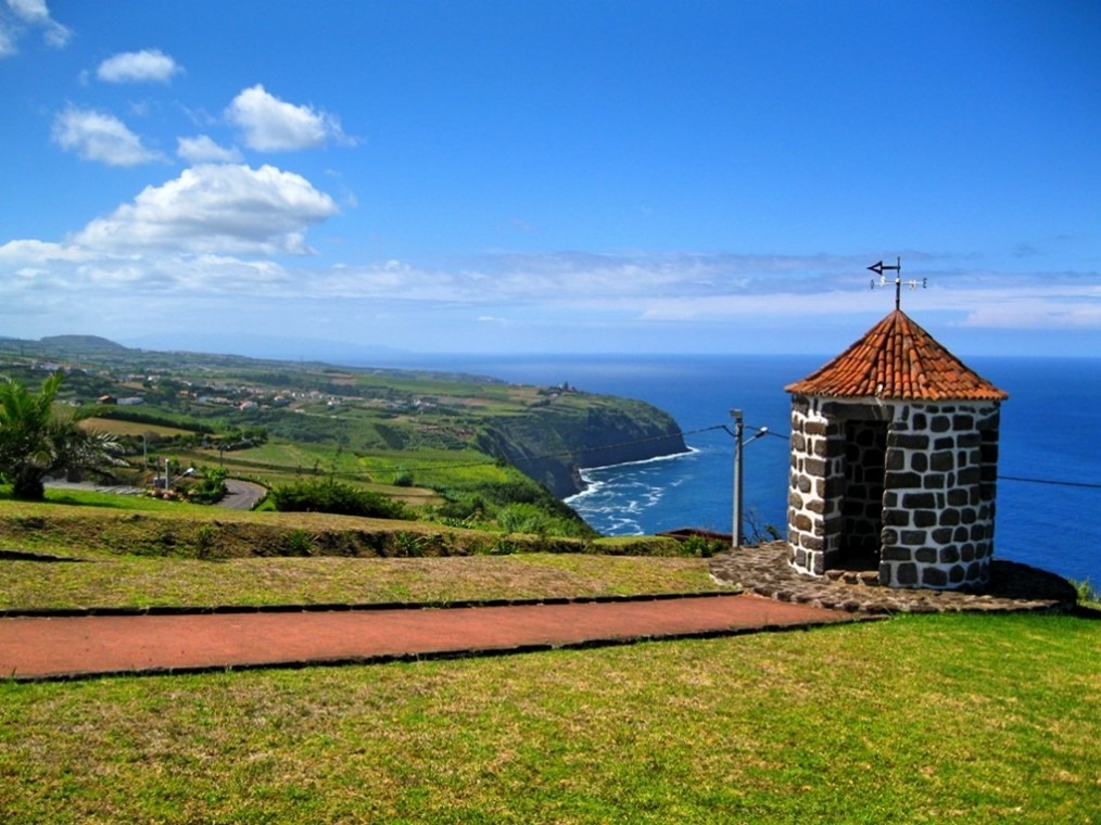 Stone gazebo by the sea on a grassy cliff with coastal view and blue sky.