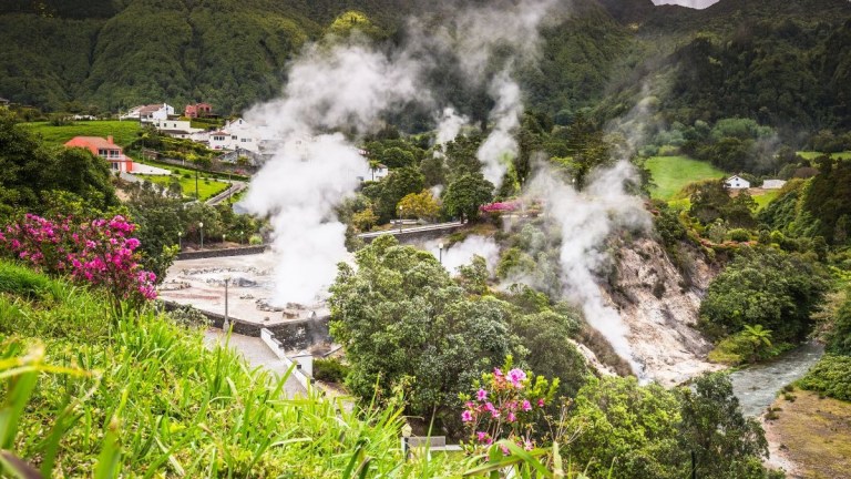 Steam rising from geothermal area with houses and greenery in the background.