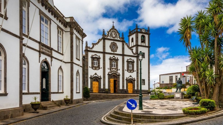 Historic stone church with ornate facade on a clear day, surrounded by palm trees and cobblestone street.
