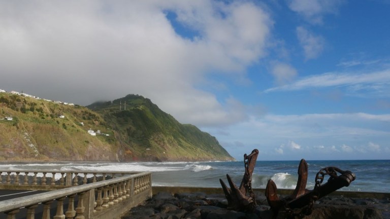 Coastal view with ocean waves, a mountain, and an anchor sculpture on a rocky shore.