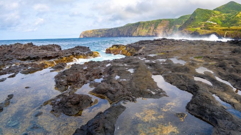 Rocky shoreline with tidal pools and distant cliffs under a cloudy sky.
