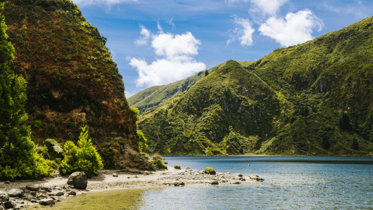 Lush green hills and a rocky shore by a calm lake under a blue sky with clouds.