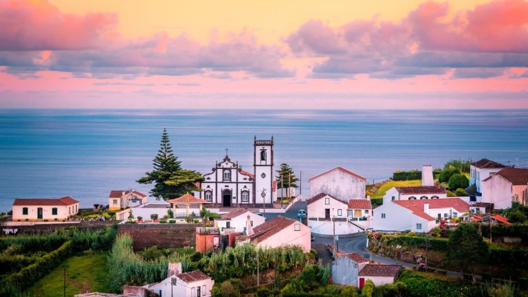 Coastal village with church, houses, and trees under a colorful sky.