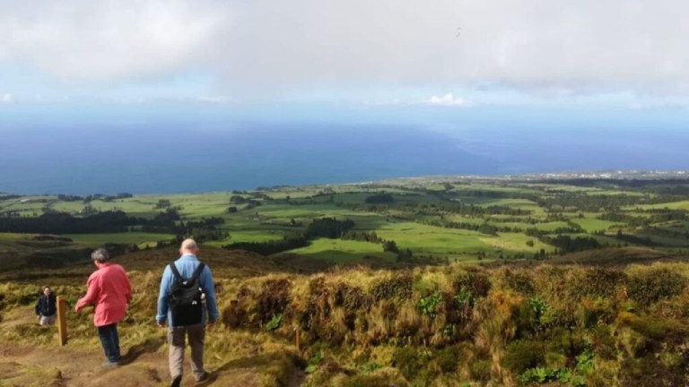 Two people walk down a grassy hill toward a vast landscape and ocean under a cloudy sky.