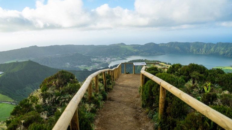 A path with wooden railings leads to a scenic lake vista in a hilly landscape.