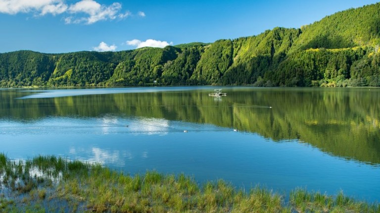 Lake with lush green hills and reflections under blue sky with clouds.