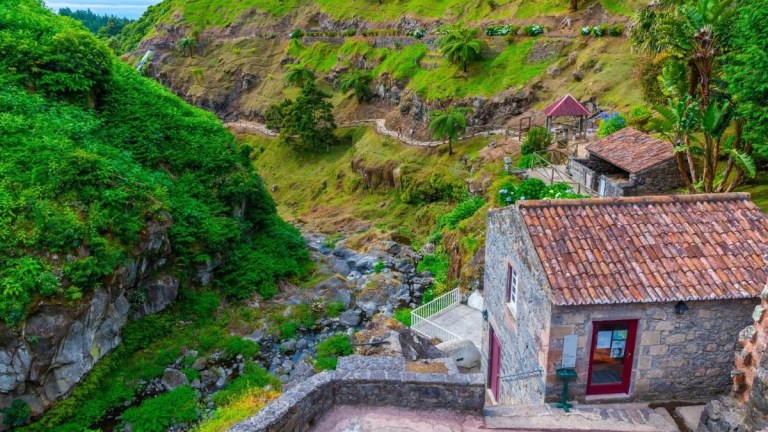 Stone house with red roof near lush green hillside and rocky stream.