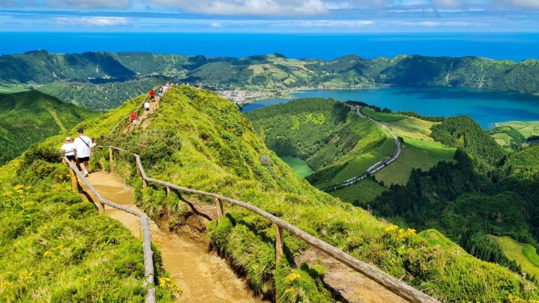 People hiking on a narrow, grassy ridge with a lake and hills in the background.