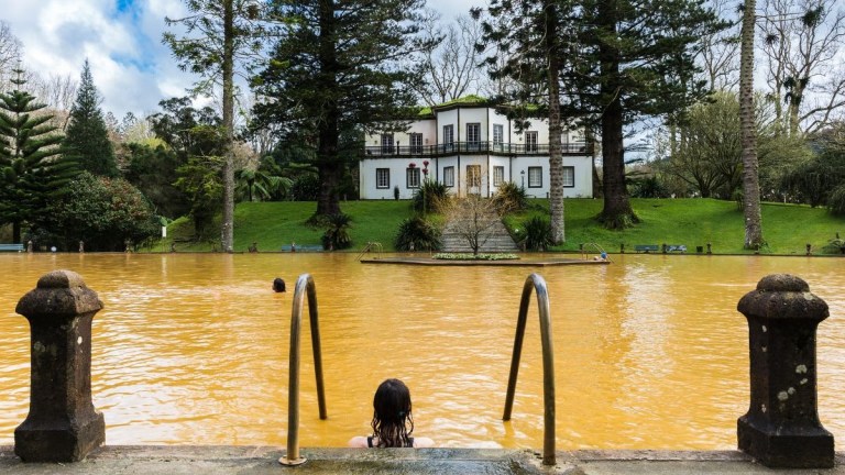 Person in orange natural pool with mansion and tall trees in the background.